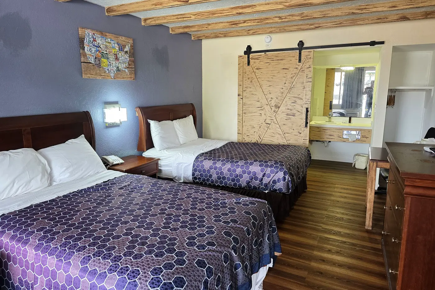 Two beds beneath timber beams with the barn door slid open toward the vanity